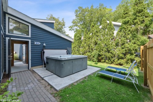 A backyard with a hot tub, lounge chairs, and greenery, featuring a blue house and a stone pathway.