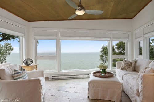 Bright living room with white furniture, a ceiling fan, and large windows overlooking a serene ocean view.