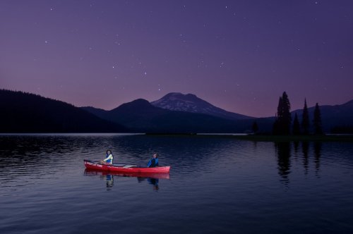 Two people in a red canoe on a calm lake at dusk, surrounded by mountains and a starry sky.