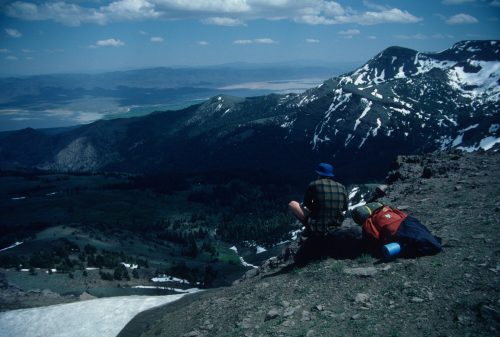 A hiker sits on a rocky ledge, overlooking a vast mountainous landscape with snow-capped peaks and a clear blue sky.
