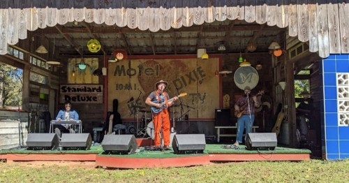 A band performs on a rustic outdoor stage with colorful decor and instruments, surrounded by a lively atmosphere.