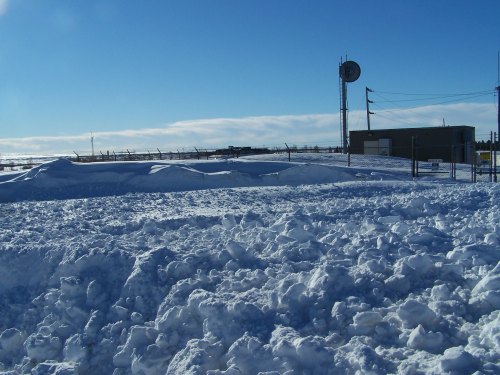 A snowy landscape with large snowdrifts under a clear blue sky, featuring a building and communication tower in the background.