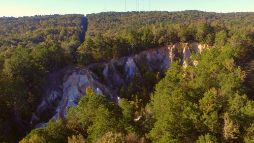 Aerial view of a rocky cliff surrounded by dense green forest under a clear blue sky.