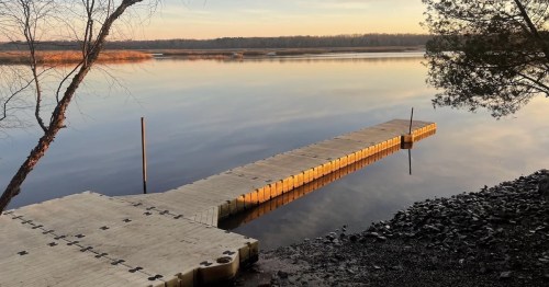 A serene lakeside view at sunset, featuring a wooden dock extending into calm waters surrounded by trees.