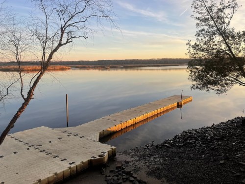 A serene lakeside view at sunset, featuring a wooden dock extending into calm waters and trees along the shore.