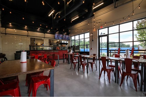 Modern restaurant interior with red chairs, wooden tables, and large windows, featuring a counter and menu screens.