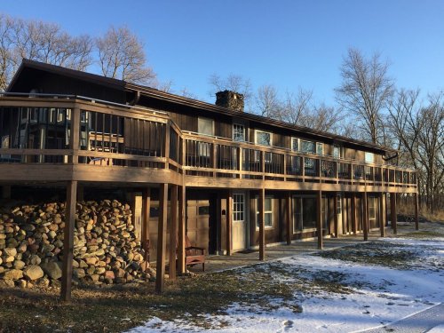 A large wooden house with a wraparound deck, surrounded by trees and a pile of stones, on a snowy ground.