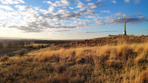 A grassy hill with a monument on top, under a blue sky with scattered clouds, overlooking a scenic landscape.
