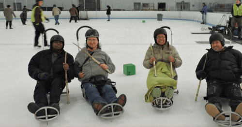 Four people sit on sleds on an ice rink, wearing helmets and holding poles, ready for a fun activity.