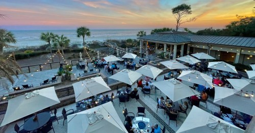 A seaside restaurant at sunset, with outdoor seating under umbrellas and guests enjoying the view.
