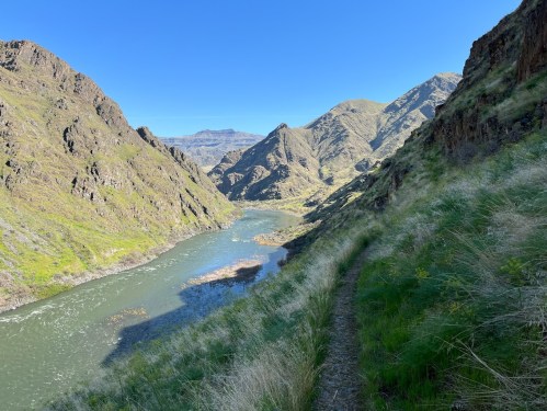 A winding river flows through a lush green canyon surrounded by steep, rocky mountains under a clear blue sky.
