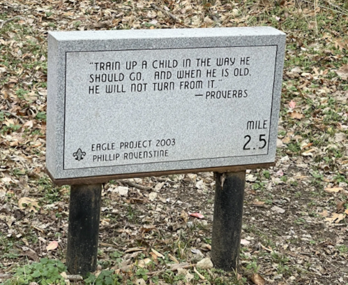 A granite mile marker with a quote from Proverbs about guiding children, placed in a natural setting.