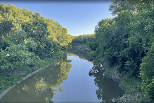 A serene river flows through a lush green landscape, reflecting trees on its calm surface under a clear sky.