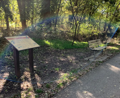 A park scene featuring an informational sign and a wooden bench surrounded by trees and greenery.