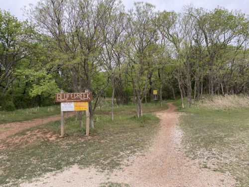A wooden sign points to Bluff Creek, with a dirt path leading into a green, wooded area.