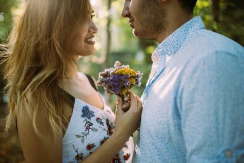 A couple stands close together in a forest, smiling and holding a bouquet of wildflowers. Soft sunlight filters through the trees.