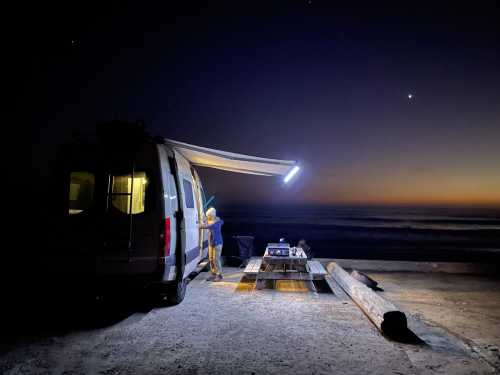 A camper van parked by the beach at dusk, with a person setting up under an illuminated awning.