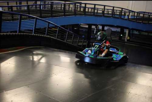 A go-kart racer navigates a curved track in an indoor racing facility, wearing a helmet and racing gear.