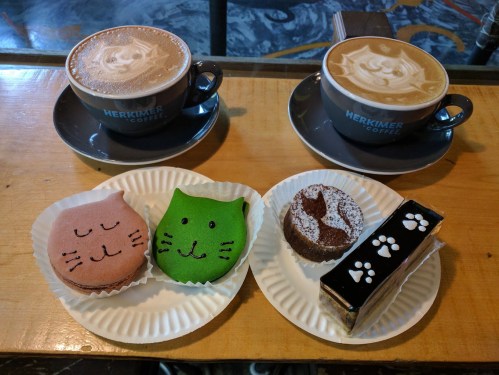 Two cups of latte art coffee beside a plate of cute, colorful cat-shaped pastries and a chocolate dessert.