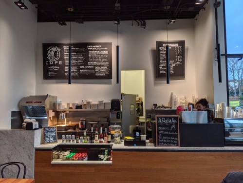 Interior of a café featuring a counter with menu boards, coffee equipment, and a barista preparing drinks.