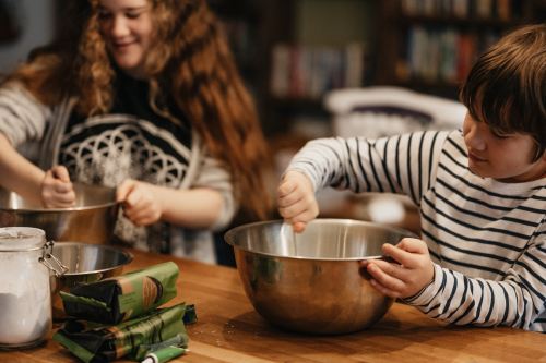 Two children mix ingredients in metal bowls on a wooden table, with baking supplies nearby.