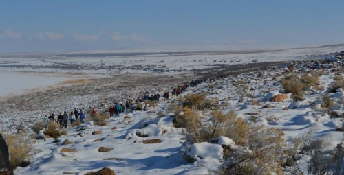 A long line of hikers traverses a snowy landscape with rocky terrain and sparse vegetation under a clear blue sky.