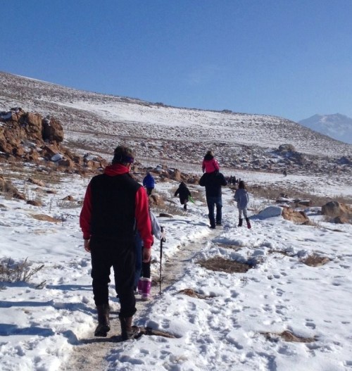 A group of people hiking on a snowy trail with rocky terrain and a clear blue sky in the background.