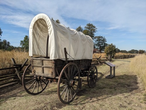 A wooden covered wagon on a grassy path, surrounded by tall grass and a wooden fence under a blue sky.