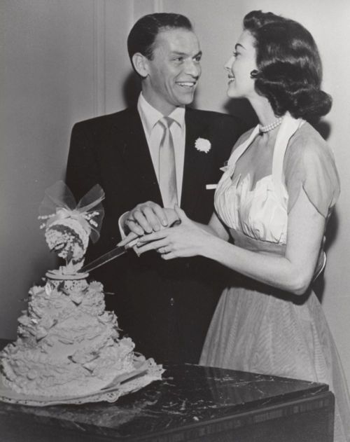 A smiling couple cuts a wedding cake together, dressed elegantly in vintage attire.