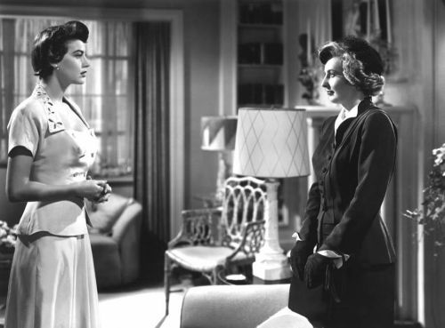Two women in vintage attire stand facing each other in a stylish, mid-century living room.