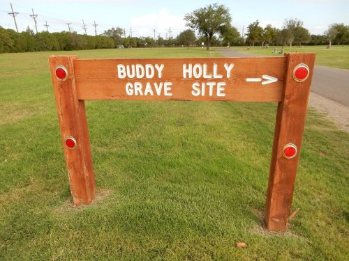 Wooden sign pointing to the Buddy Holly grave site, with red reflectors, set in a grassy area.