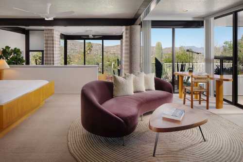 Modern bedroom with a curved sofa, wooden table, and large windows showcasing a desert view. Bright and airy atmosphere.