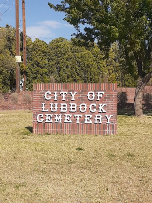 Sign for the City of Lubbock Cemetery, displayed on a brick structure with green grass and trees in the background.