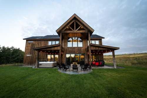 A modern wooden house with large windows, a patio, and outdoor seating, set in a grassy landscape under a cloudy sky.