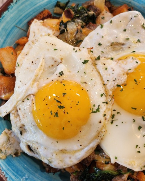 A close-up of a blue plate with two sunny-side-up eggs on a bed of sautéed potatoes and greens, garnished with herbs.