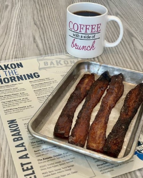 A tray of crispy bacon strips next to a coffee mug and a brunch menu on a wooden table.
