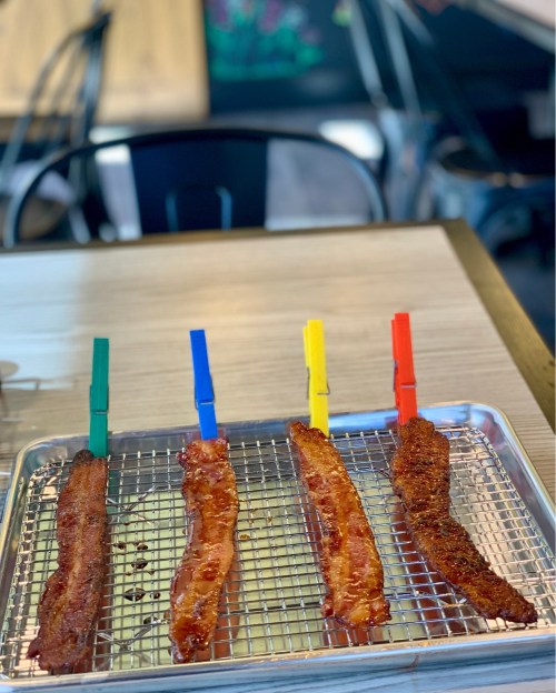 Four strips of bacon on a wire rack, each secured with a colorful clothespin, on a wooden table.
