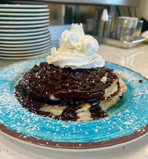 A stack of pancakes topped with chocolate sauce, mini chocolate chips, and whipped cream, served on a blue plate.