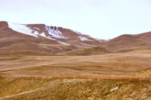 A vast landscape of rolling hills with sparse vegetation and a dusting of snow on the peaks in the background.