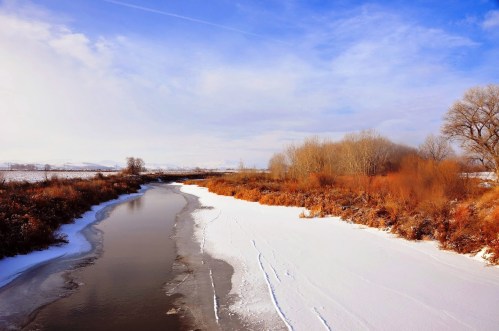 A serene winter landscape featuring a river bordered by snow and bare trees under a blue sky.