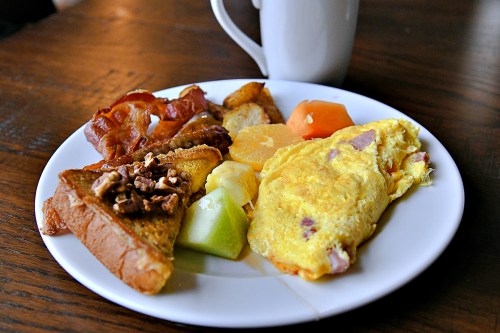 A plate of breakfast featuring an omelet, bacon, toast with nuts, fruit, and a cup of coffee in the background.