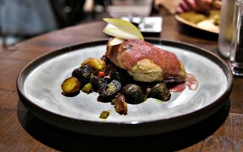 A plate of roasted chicken topped with a berry sauce, served with colorful roasted vegetables and a slice of apple.