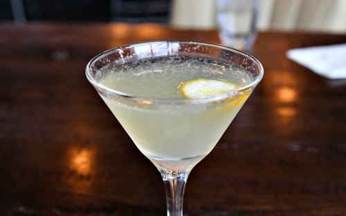 A close-up of a cocktail in a martini glass, garnished with a lemon slice, on a wooden table.