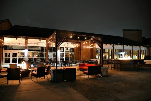 Outdoor seating area at night with string lights, fire pit, and modern furniture near a building.