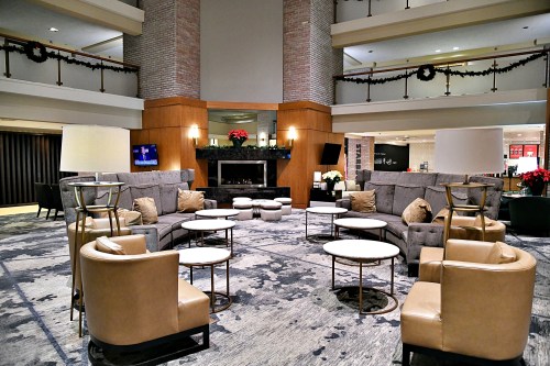 Modern hotel lobby with cozy seating, circular tables, and festive decorations, featuring a fireplace and TV.