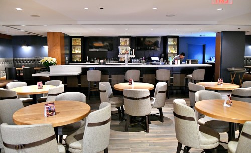 A modern bar area with round wooden tables and a sleek counter, featuring a bartender in the background.