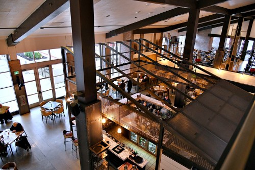 A spacious restaurant interior with high ceilings, large windows, and diners seated at tables.