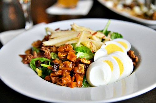 A fresh salad with chopped greens, sliced apples, grilled chicken, crispy bacon, and hard-boiled eggs on a white plate.