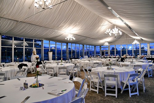Elegant event space with round tables, white tablecloths, and chandeliers under a draped ceiling, set for a gathering.