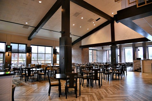 Modern restaurant interior with wooden tables, chairs, and large windows, featuring high ceilings and a spacious layout.